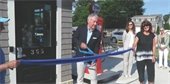 Man in blue blazer cutting a blue ribbon with large scissors in front of a small building with an old fashioned red gas pump next to it