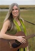 Woman with long blonde hair holding an acoustic guitar standing in front of a saltmarsh