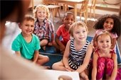 a group of preschoolers seated on the floor of a classroom smiling at someone off camera