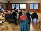 A group of people seated in folding chairs with their backs to the camera. A screen and projector are in the front of the room.