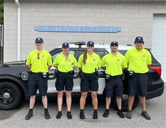 Five young people in dark shorts and hi-vis shirts standing in front of a police cruiser