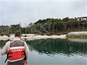 Man in USFWS life vest stands in a boat looking at dead trees on the shore