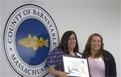 Two women standing in front of the Barnstable County seal holding a certificate.