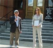 A man holding a microphone and a woman holding a certificate in from of a stone and brick library