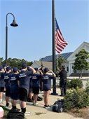 Young boys and girls wearing navy T-shirts with Youth Academy on the back saluting the American flag as it is raised on a flag pole.