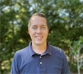 A man in a dark blue golf shirt with short brown hair standing outdoors with green trees in the background