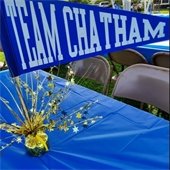 Blue pennant with Team Chatham spelled out in white letter on a wooden dowel stuck in a spray of gold foil ona table covered with a blue plastic tablecloth