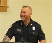 Man with close cropped hair standing at a podium in a blue uniform shirt smiling