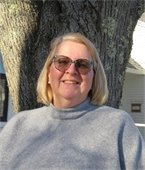 A woman with short blond hair wearing a grey sweater standing in front of a tree