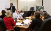 Men and women around a conference table watching a man write on a white board