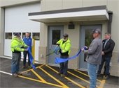 Five men standing in front of an industrial building. Two are holding the ends of a blue ribbon while a third cuts through it using oversized scissors