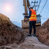 Person in orange safety vest standing on a pipe in the ground in front of an excavator