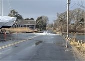 A white car driving through a flooded street with a salt marsh on one side and boats on the other