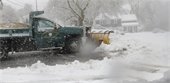 Green pick up truck with sand in the bed and a yellow plow on it plowing snow off a street